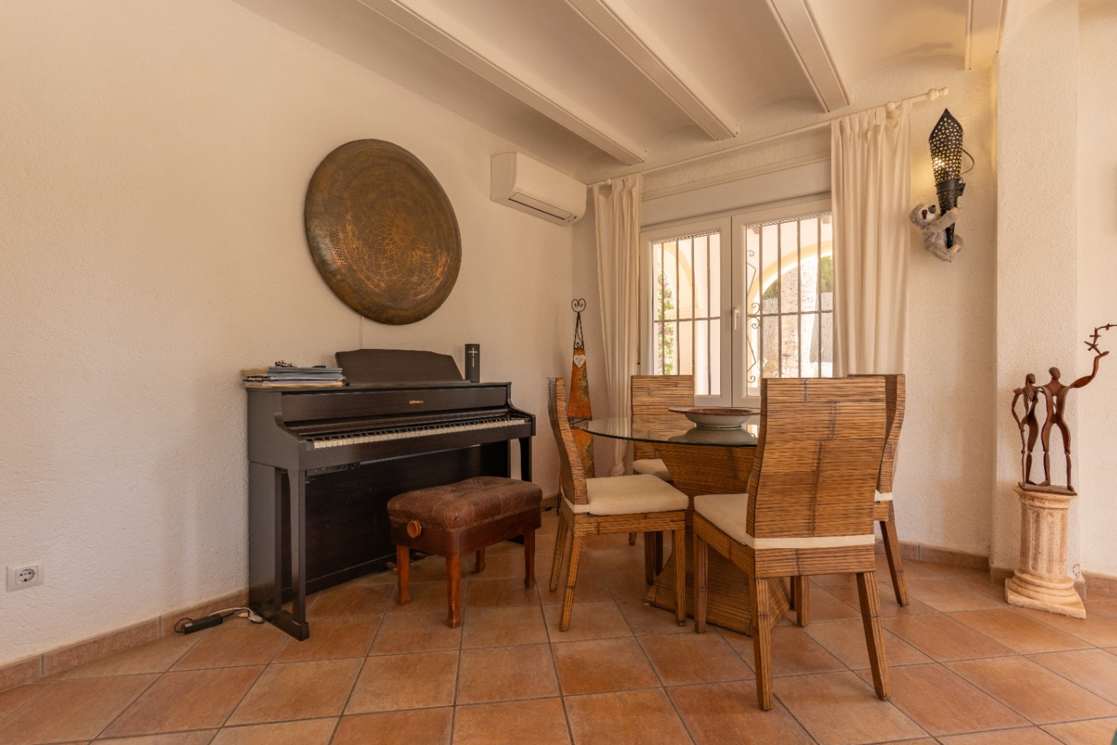 Coin repas avec carrelage couleur terre cuite, piano numérique et table en verre entourée de chaises en bambou. La fenêtre à double vitrage et barreaux apporte une lumière naturelle.