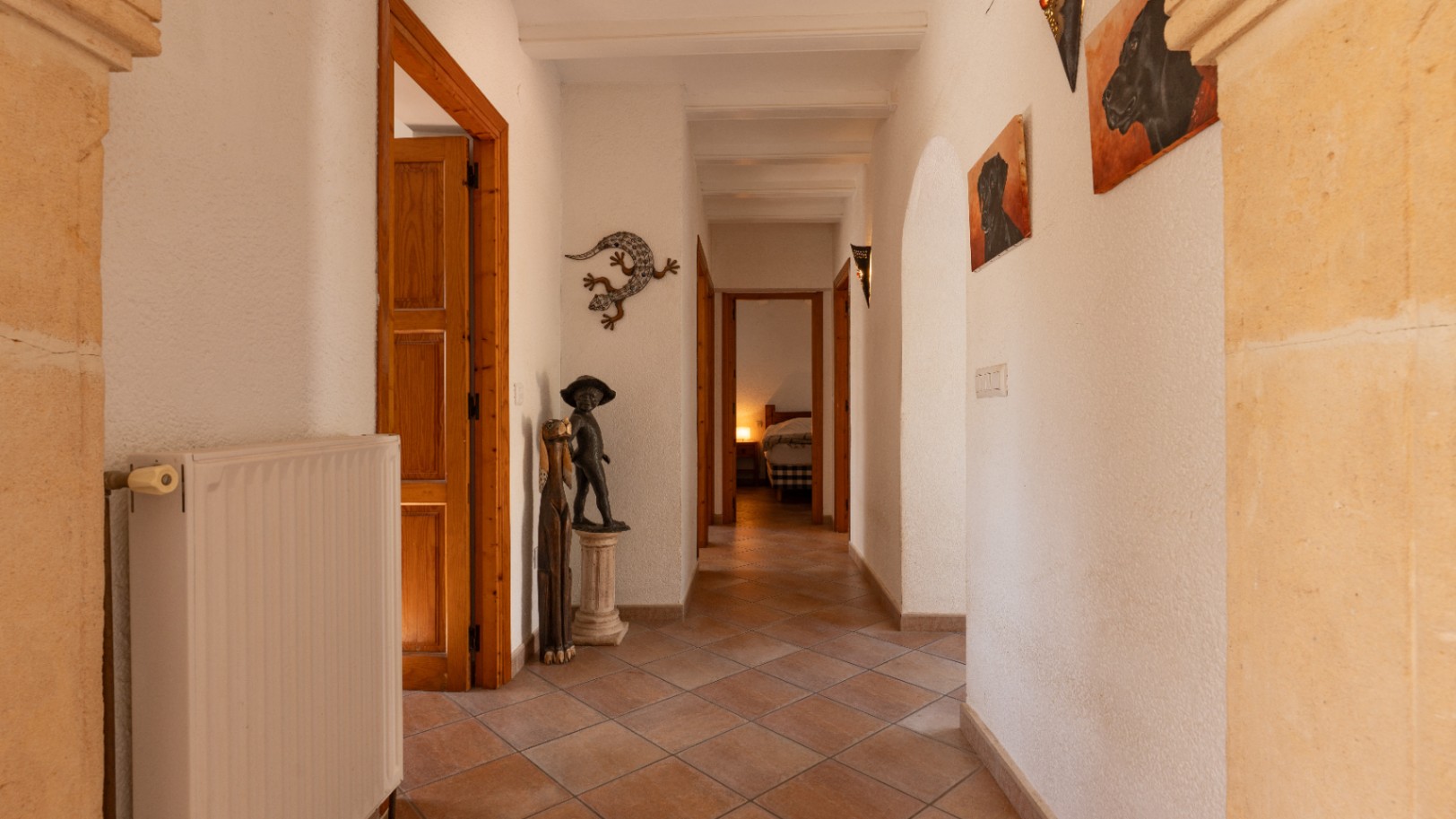 Couloir lumineux avec sol en carreaux de terre cuite, murs blancs texturés et poutres en bois apparentes. On y trouve un radiateur, des statues décoratives et une vue sur une chambre.