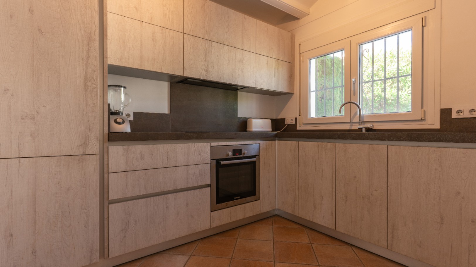 Cuisine rénovée en L avec des armoires effet bois clair, des plans de travail gris foncé et un four intégré. Une fenêtre PVC blanche avec barreaux offre de la lumière naturelle.
