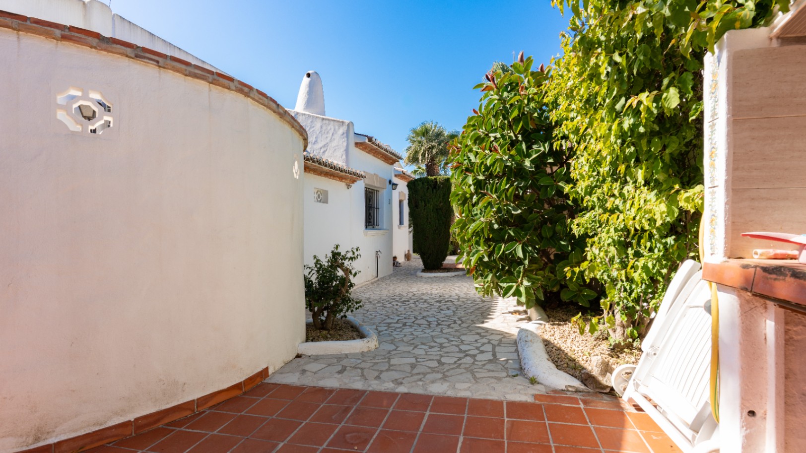 Entrée de la propriété avec un chemin en pierres irrégulières, bordé d'un mur blanc incurvé avec un motif ajouré et une végétation méditerranéenne luxuriante. Carrelage en terre cuite au premier plan.