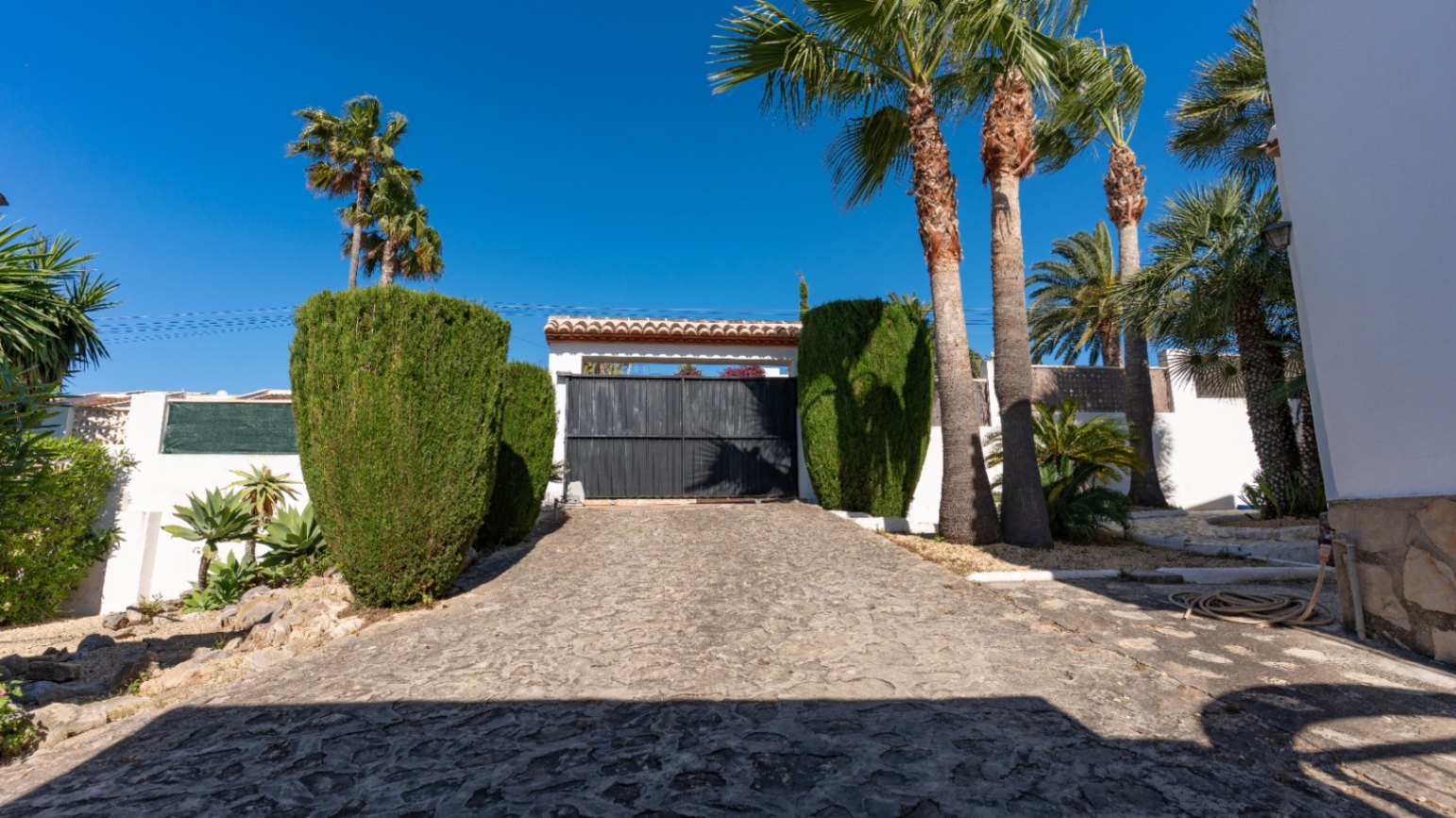 Entrée principale avec portail automatique sombre et allée pavée, bordée de palmiers et de haies vertes sous un ciel azur. Une toiture en tuiles traditionnelles surmonte l'accès.