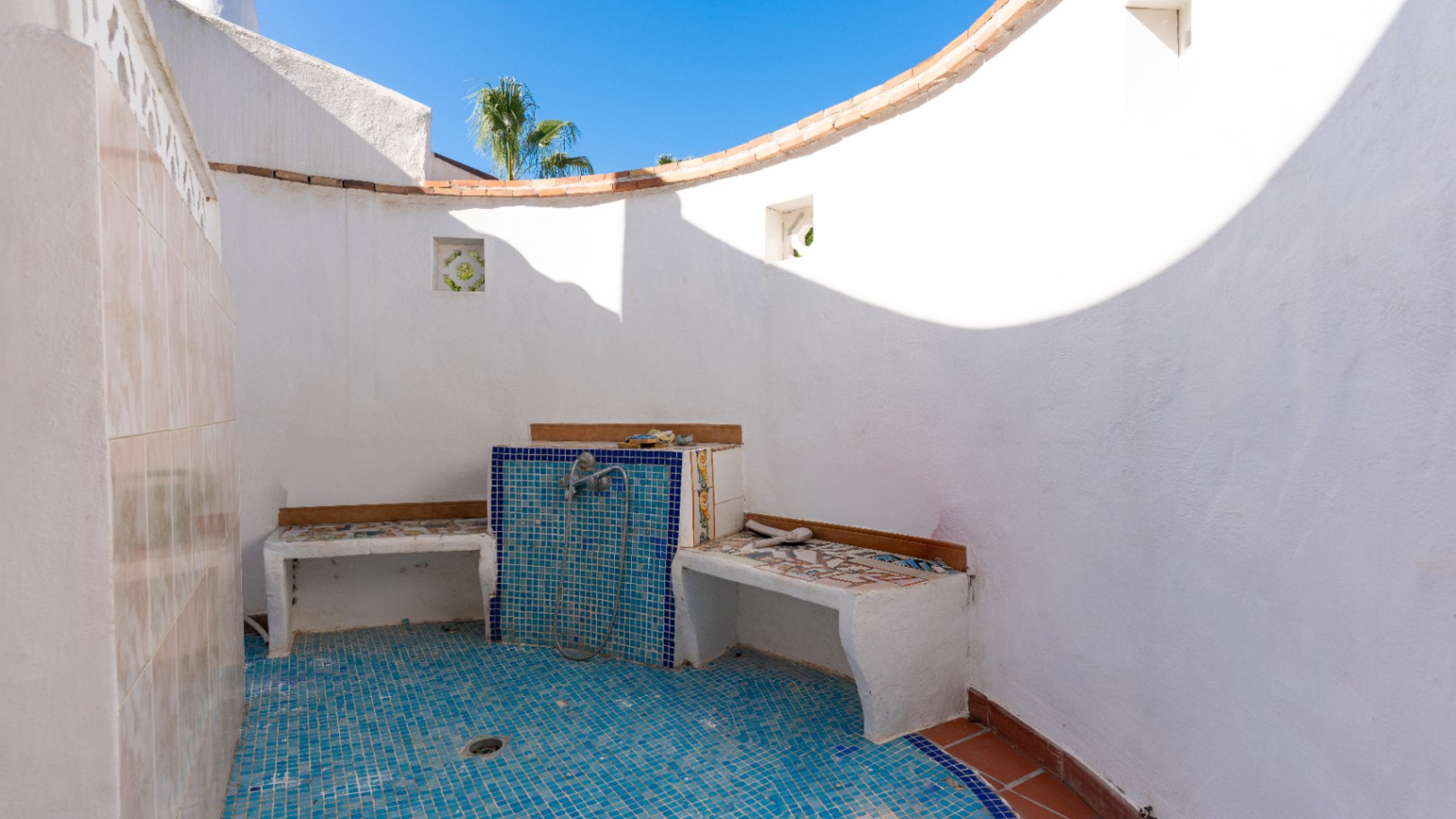 Espace douche extérieur avec murs blancs crépis, sol en mosaïque bleue et bancs carrelés intégrés. Un mur incurvé s'ouvre sur le ciel bleu.