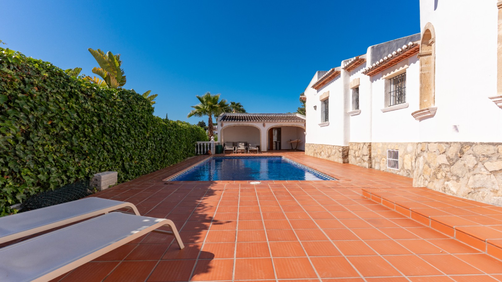 Espace piscine privé et ensoleillé avec piscine rectangulaire, terrasse en terre cuite et deux chaises longues. Une haie verte dense assure l'intimité près de la villa blanche.