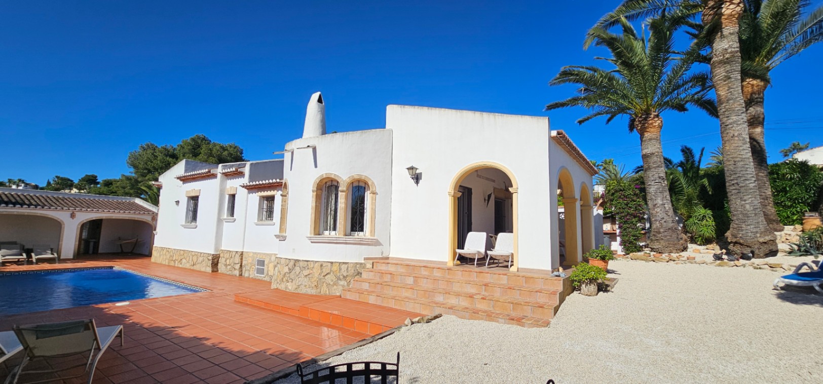 Extérieur de villa blanche avec soubassement en pierre, escaliers en terre cuite et porche voûté. Piscine avec terrasse carrelée et jardin méditerranéen avec palmiers sous ciel bleu.
