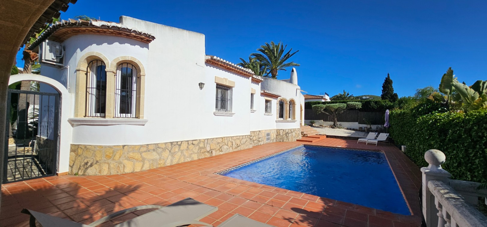 Extérieur de villa blanche avec soubassement en pierre et fenêtres cintrées grillagées. Piscine rectangulaire bordée de carrelage en terre cuite, palmiers et verdure sous un ciel azur.