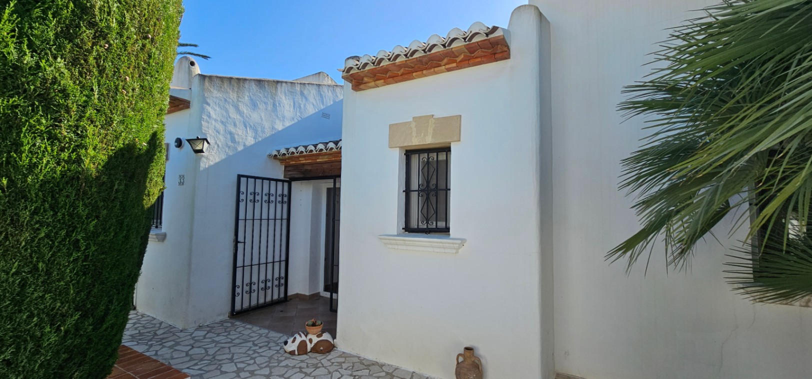Extérieur de villa blanche avec tuiles en terre cuite, fenêtre à barreaux en fer forgé décoratifs et linteau en pierre. L'entrée s'ouvre sur un portail en fer et une haie verte.