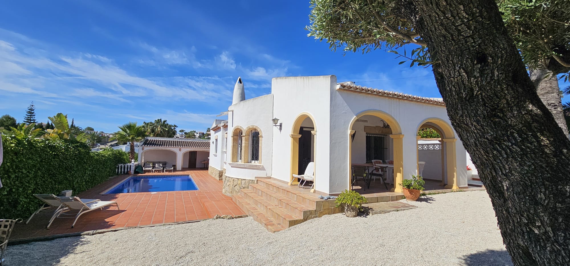 Extérieur de villa méditerranéenne blanche avec des arcs jaunes. La piscine carrelée en terre cuite et une terrasse couverte pour les repas extérieurs sont présentes.