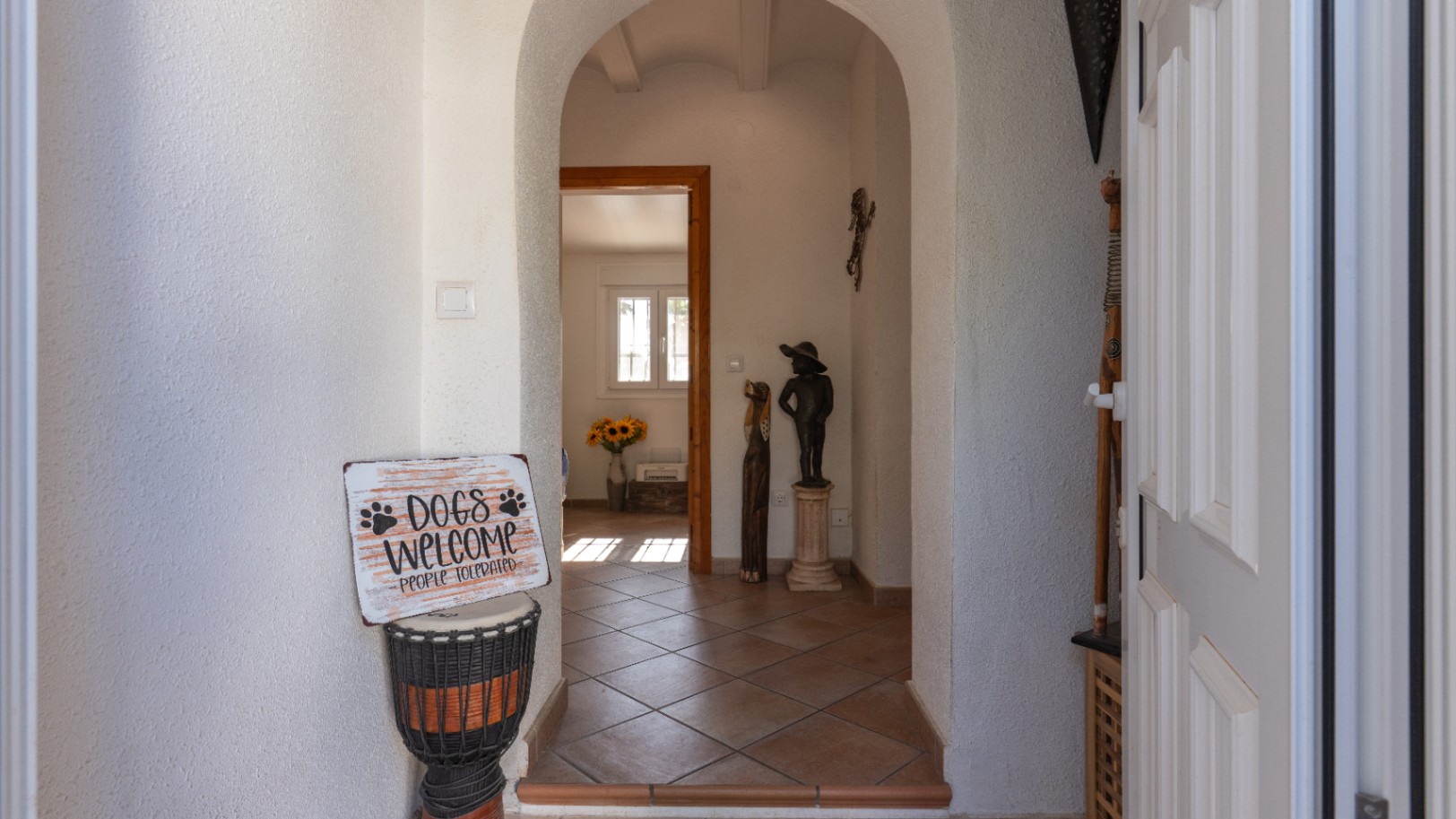 Hall d'entrée avec des arches blanches et un sol carrelé en terre cuite. Une porte d'entrée à panneaux est visible à droite, menant à une pièce lumineuse avec des tournesols.