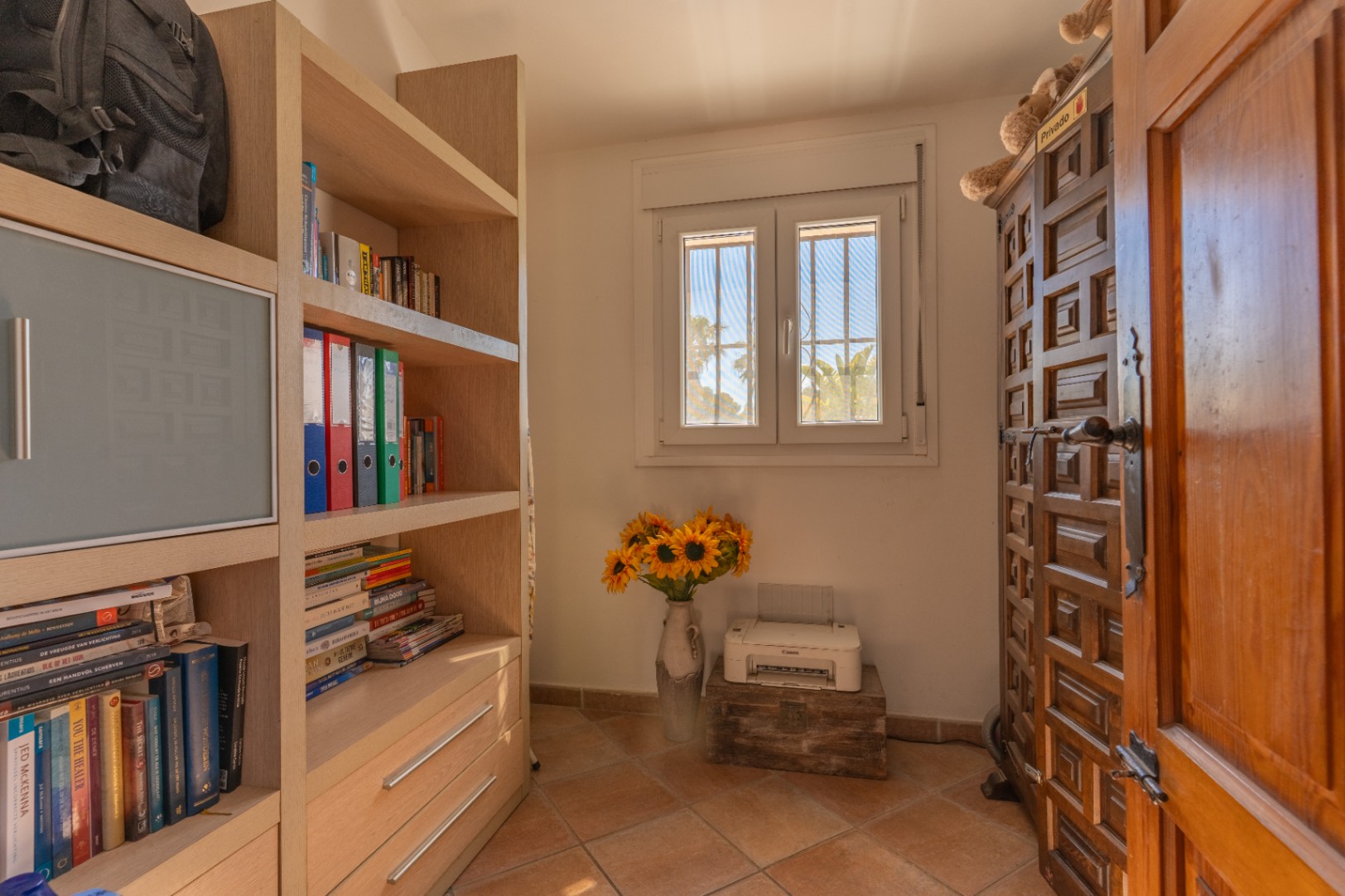 Pièce avec une étagère en bois clair, une fenêtre PVC à double vitrage et barreaux, offrant une vue sur les palmiers. Porte en bois sculpté et sol carrelé.