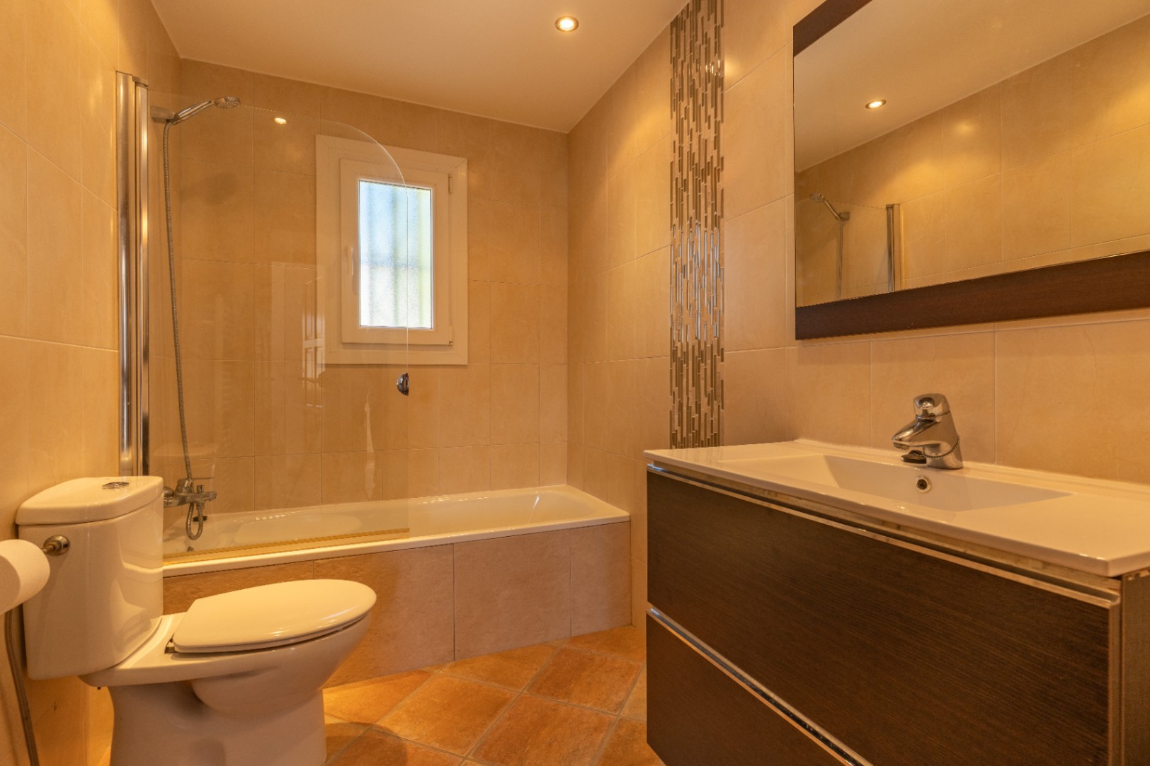 Salle de bain lumineuse avec baignoire et paroi vitrée, meuble-lavabo aspect bois foncé avec vasque blanche, et carrelage mural beige avec bande mosaïque décorative.