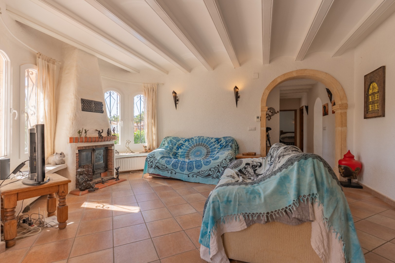 Salon lumineux avec cheminée traditionnelle en briques, fenêtres cintrées à barreaux et plafond à poutres apparentes. Le sol carrelé de terre cuite et une arche en pierre mènent à l'intérieur.