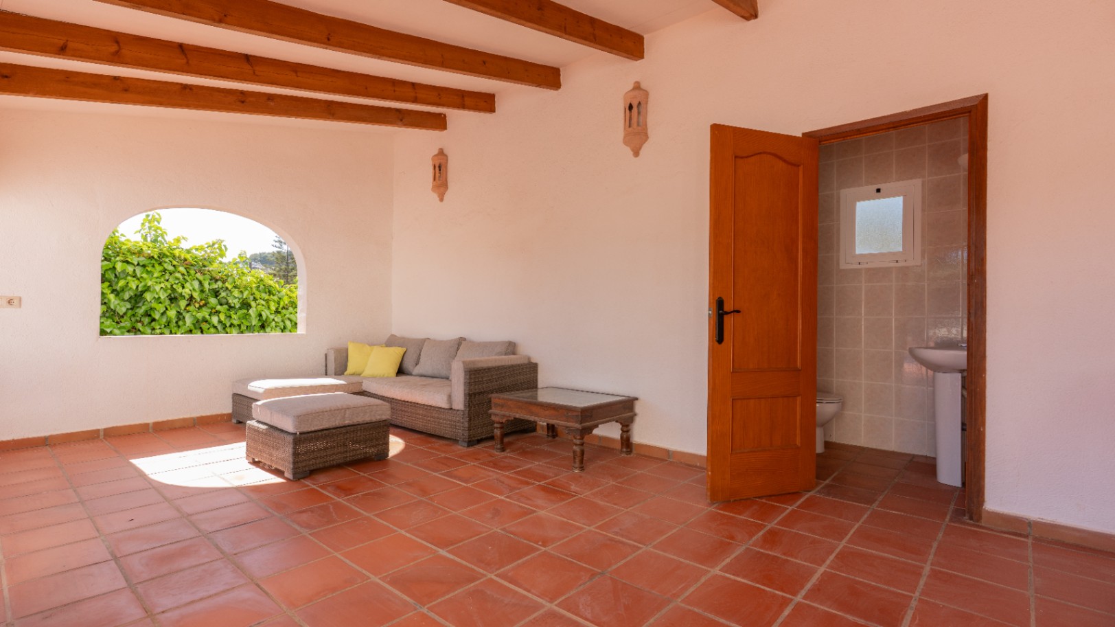 Terrasse couverte avec sol en terre cuite, poutres apparentes et canapé en rotin. Une fenêtre arquée donne sur la verdure, avec un accès attenant à des toilettes.