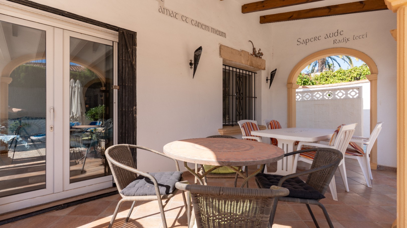 Terrasse couverte avec sol en terre cuite, table ronde en mosaïque et portes vitrées en PVC blanc. Une arche encadre la végétation luxuriante.