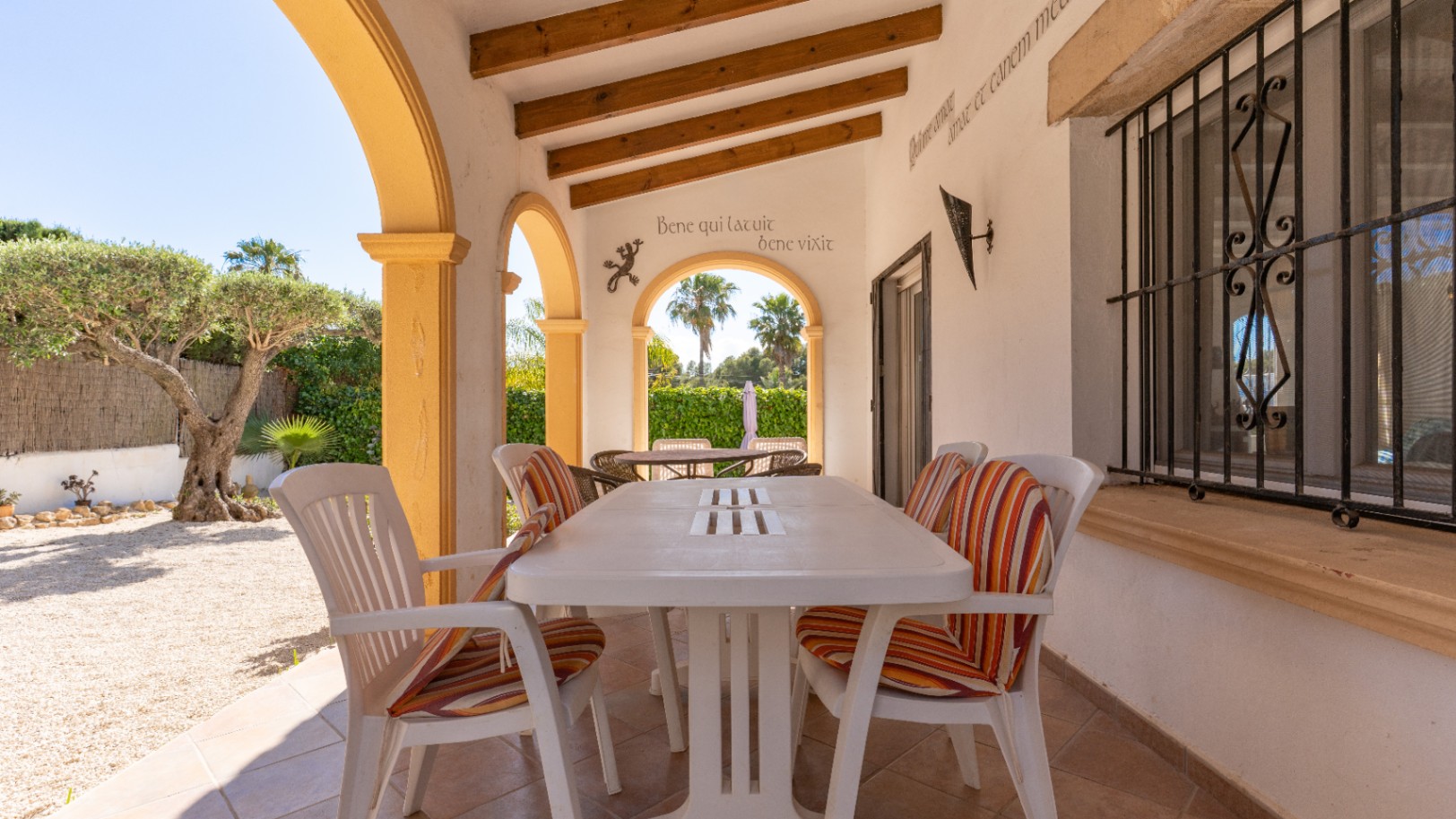 Terrasse couverte carrelée avec table à manger blanche et chaises à coussins rayés. Arches jaunes, poutres apparentes et vue sur un jardin de gravier avec olivier.