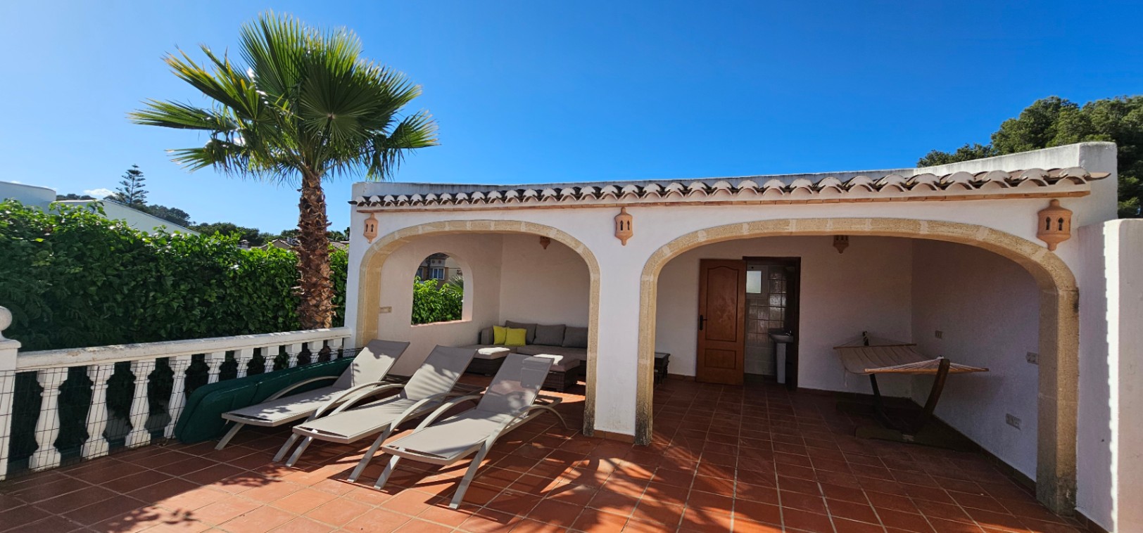 Vaste terrasse de piscine carrelée en terre cuite, trois chaises longues et une naya couverte avec salon extérieur et toilettes intégrées.