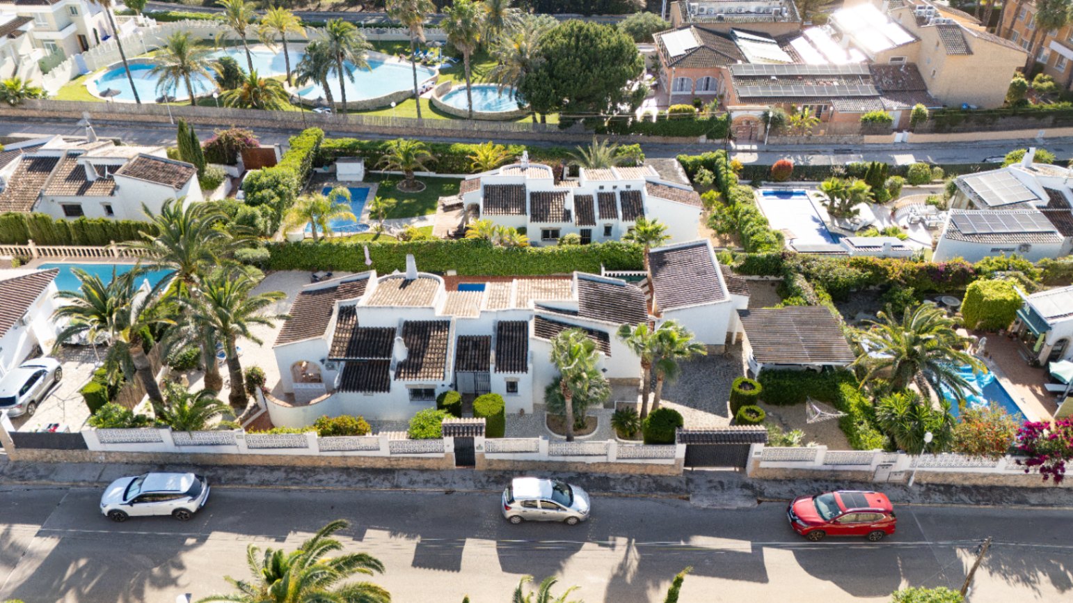 Vue aérienne de la villa privée à Javea, avec sa façade blanche, ses toits sombres, une grande piscine et un jardin méditerranéen luxuriant avec palmiers et entrée privée.