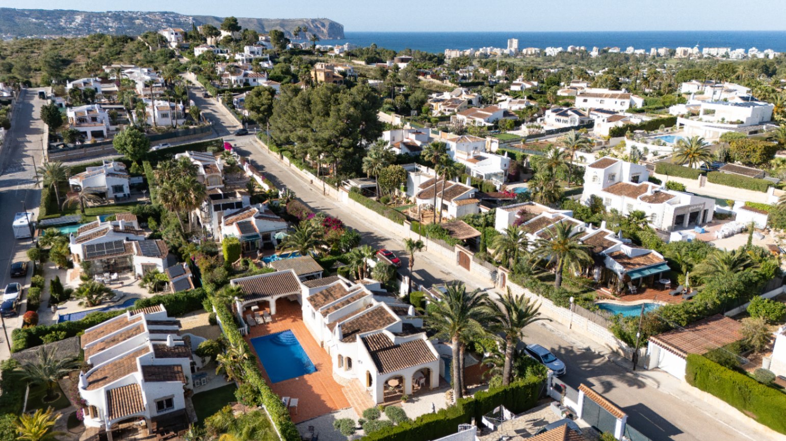 Vue aérienne du quartier résidentiel paisible de Javea, présentant de nombreuses villas blanches avec piscines privées, jardins méditerranéens luxuriants et la mer bleue au loin.