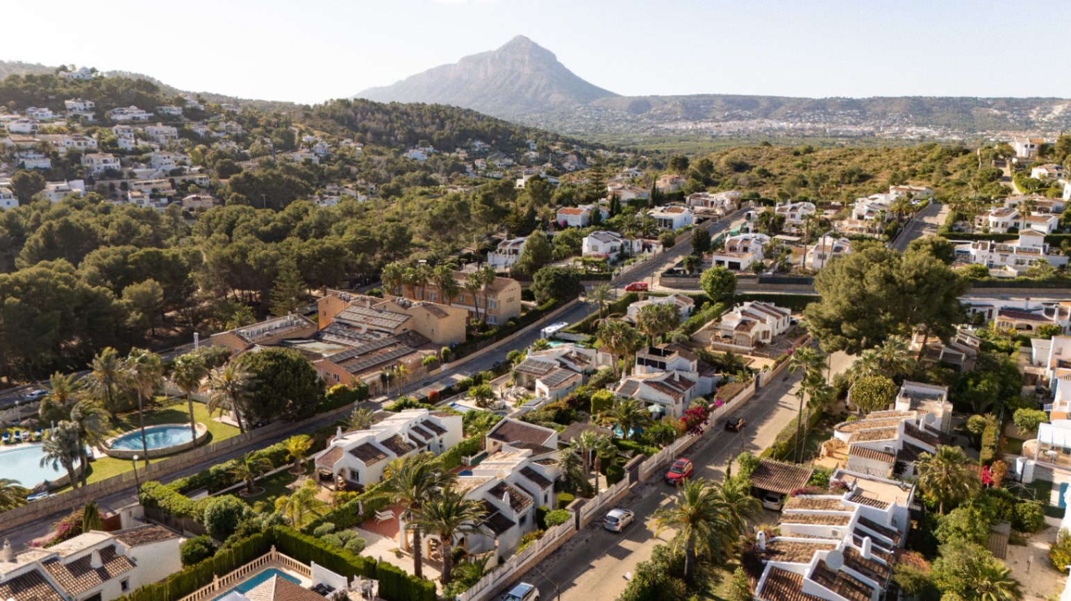 Vue aérienne d'un quartier résidentiel avec villas blanches et toits en terre cuite, entourées de végétation méditerranéenne luxuriante et de rues sinueuses, avec une montagne en arrière-plan.