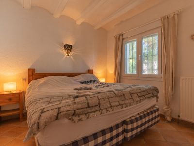 Chambre à coucher avec lit en bois et base à carreaux, sol en terre cuite et fenêtre PVC à double vitrage. Une applique murale décorative projette des motifs lumineux, complétant le plafond à poutres apparentes.