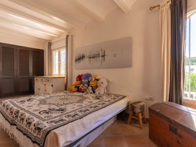 Chambre lumineuse avec sol en terre cuite, murs blancs texturés et poutres apparentes. Armoires à persiennes foncées, lit avec couverture à motifs et lumière naturelle des fenêtres PVC double vitrage.