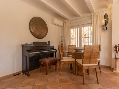 Coin repas avec carrelage couleur terre cuite, piano numérique et table en verre entourée de chaises en bambou. La fenêtre à double vitrage et barreaux apporte une lumière naturelle.