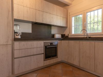 Cuisine rénovée en L avec des armoires effet bois clair, des plans de travail gris foncé et un four intégré. Une fenêtre PVC blanche avec barreaux offre de la lumière naturelle.