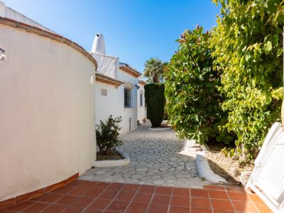 Entrée de la propriété avec un chemin en pierres irrégulières, bordé d'un mur blanc incurvé avec un motif ajouré et une végétation méditerranéenne luxuriante. Carrelage en terre cuite au premier plan.