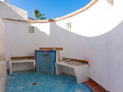 Espace douche extérieur avec murs blancs crépis, sol en mosaïque bleue et bancs carrelés intégrés. Un mur incurvé s'ouvre sur le ciel bleu.