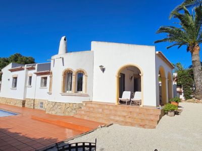 Extérieur de villa blanche avec soubassement en pierre, escaliers en terre cuite et porche voûté. Piscine avec terrasse carrelée et jardin méditerranéen avec palmiers sous ciel bleu.