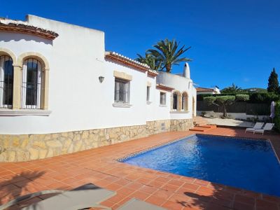 Extérieur de villa blanche avec soubassement en pierre et fenêtres cintrées grillagées. Piscine rectangulaire bordée de carrelage en terre cuite, palmiers et verdure sous un ciel azur.