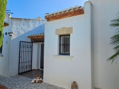 Extérieur de villa blanche avec tuiles en terre cuite, fenêtre à barreaux en fer forgé décoratifs et linteau en pierre. L'entrée s'ouvre sur un portail en fer et une haie verte.