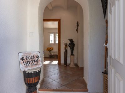 Hall d'entrée avec des arches blanches et un sol carrelé en terre cuite. Une porte d'entrée à panneaux est visible à droite, menant à une pièce lumineuse avec des tournesols.