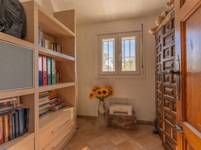 Pièce avec une étagère en bois clair, une fenêtre PVC à double vitrage et barreaux, offrant une vue sur les palmiers. Porte en bois sculpté et sol carrelé.