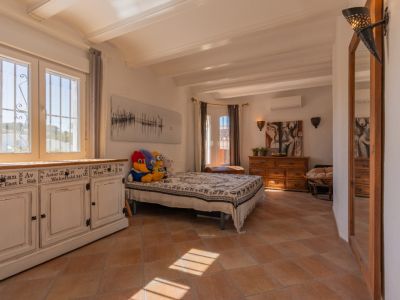Pièce lumineuse avec carrelage en terre cuite et poutres blanches. Un meuble blanc patiné se trouve près de fenêtres PVC avec barreaux, inondée de lumière naturelle.
