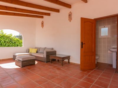 Terrasse couverte avec sol en terre cuite, poutres apparentes et canapé en rotin. Une fenêtre arquée donne sur la verdure, avec un accès attenant à des toilettes.