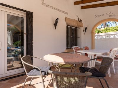 Terrasse couverte avec sol en terre cuite, table ronde en mosaïque et portes vitrées en PVC blanc. Une arche encadre la végétation luxuriante.