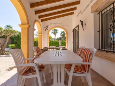 Terrasse couverte carrelée avec table à manger blanche et chaises à coussins rayés. Arches jaunes, poutres apparentes et vue sur un jardin de gravier avec olivier.
