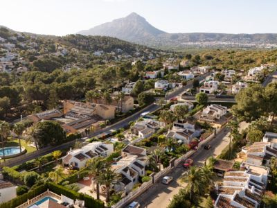 Vue aérienne d'un quartier résidentiel avec villas blanches et toits en terre cuite, entourées de végétation méditerranéenne luxuriante et de rues sinueuses, avec une montagne en arrière-plan.