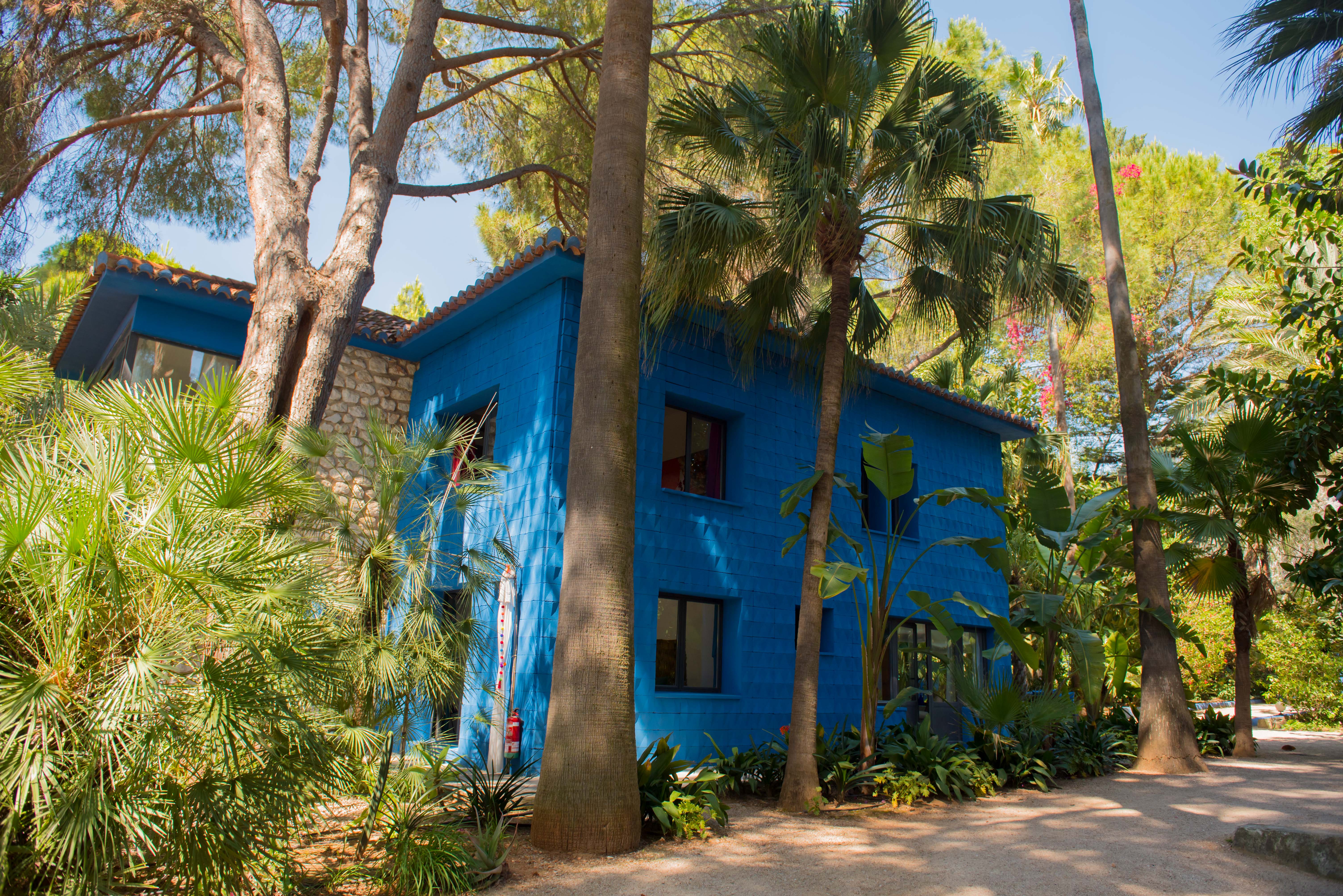 Bâtiment de deux étages à la façade bleue texturée et vibrante, avec fenêtres sombres et un mur en pierre, intégré dans un jardin tropical luxuriant traversé par un chemin de gravier.