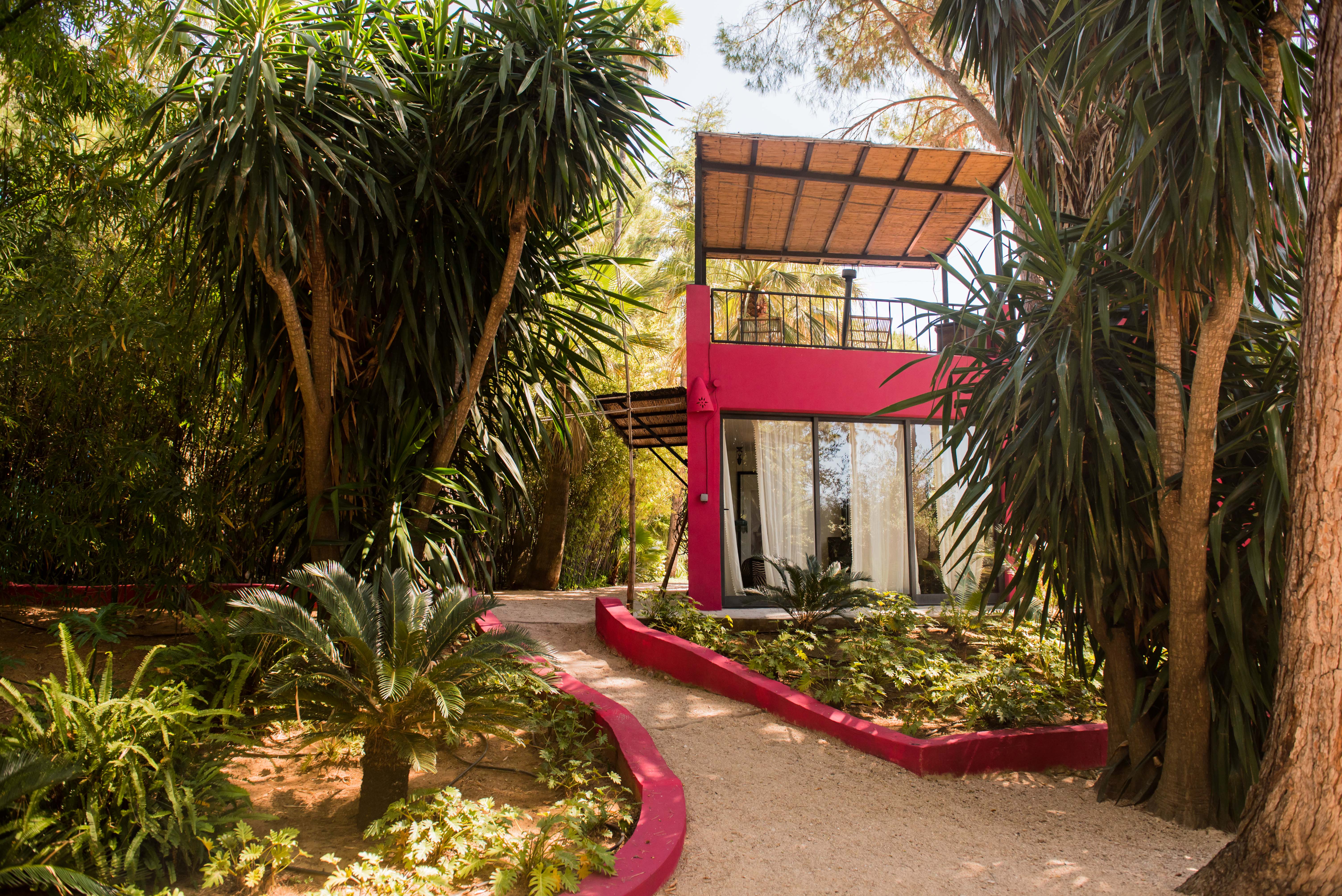 Bâtiment moderne fuchsia avec grandes portes vitrées, auvent en matériaux naturels et balcon supérieur avec sièges en rotin. Un chemin de gravier serpente à travers la végétation tropicale luxuriante.