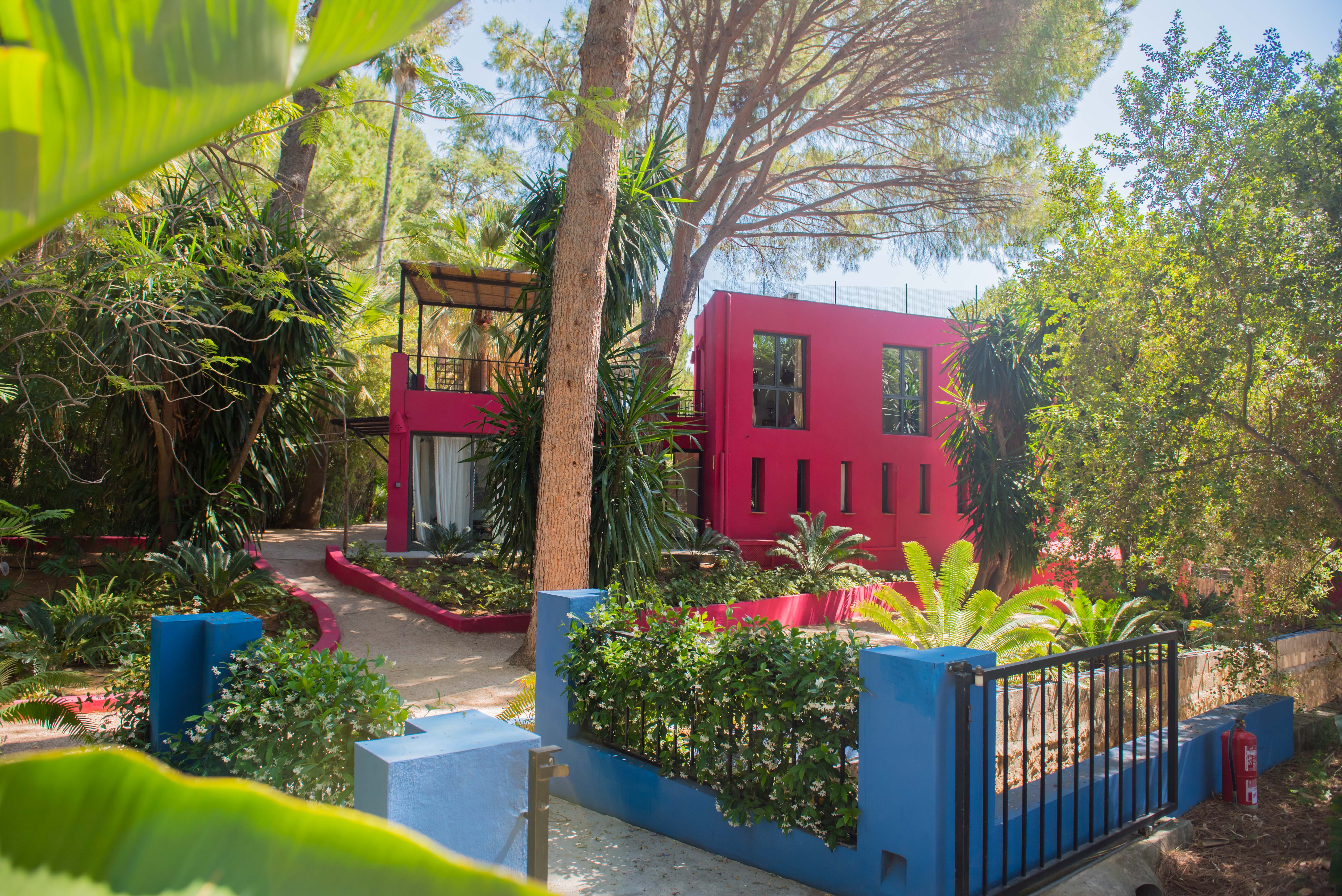 Bâtiment moderne fuchsia éclatant avec fenêtres variées, niché dans un jardin tropical dense avec de grands arbres et des sentiers sinueux. Un muret bleu fleuri délimite le premier plan.