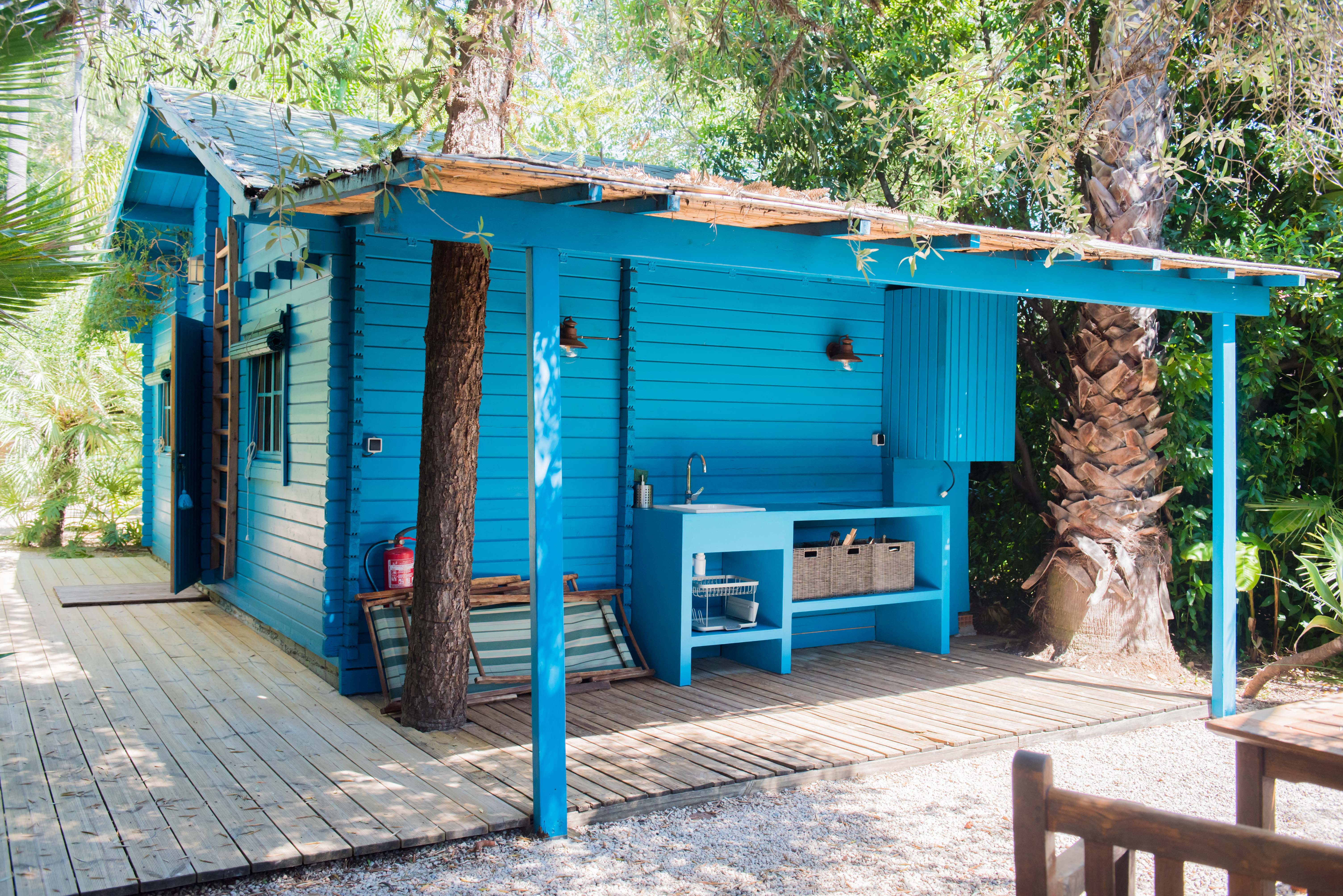 Cabane en bois bleu vif avec cuisine extérieure intégrée, évier et étagères ouvertes, sur une terrasse en bois. Des arbres tropicaux traversent sa canopée naturelle, entourée de chemins de gravier.