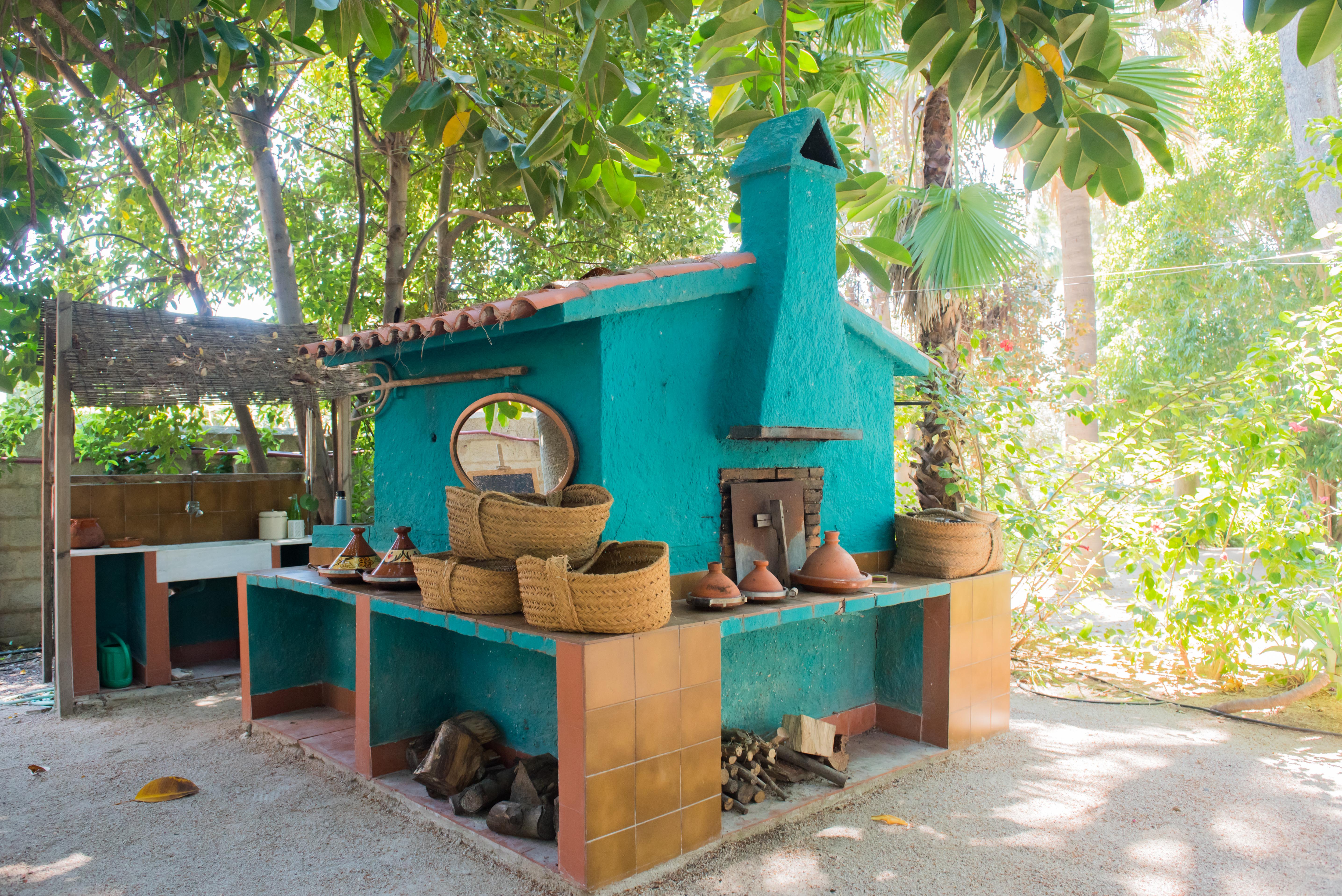 Espace de cuisson extérieur avec four turquoise et cheminée, comptoirs carrelés en terre cuite et rangement pour le bois, niché dans un jardin tropical luxuriant.