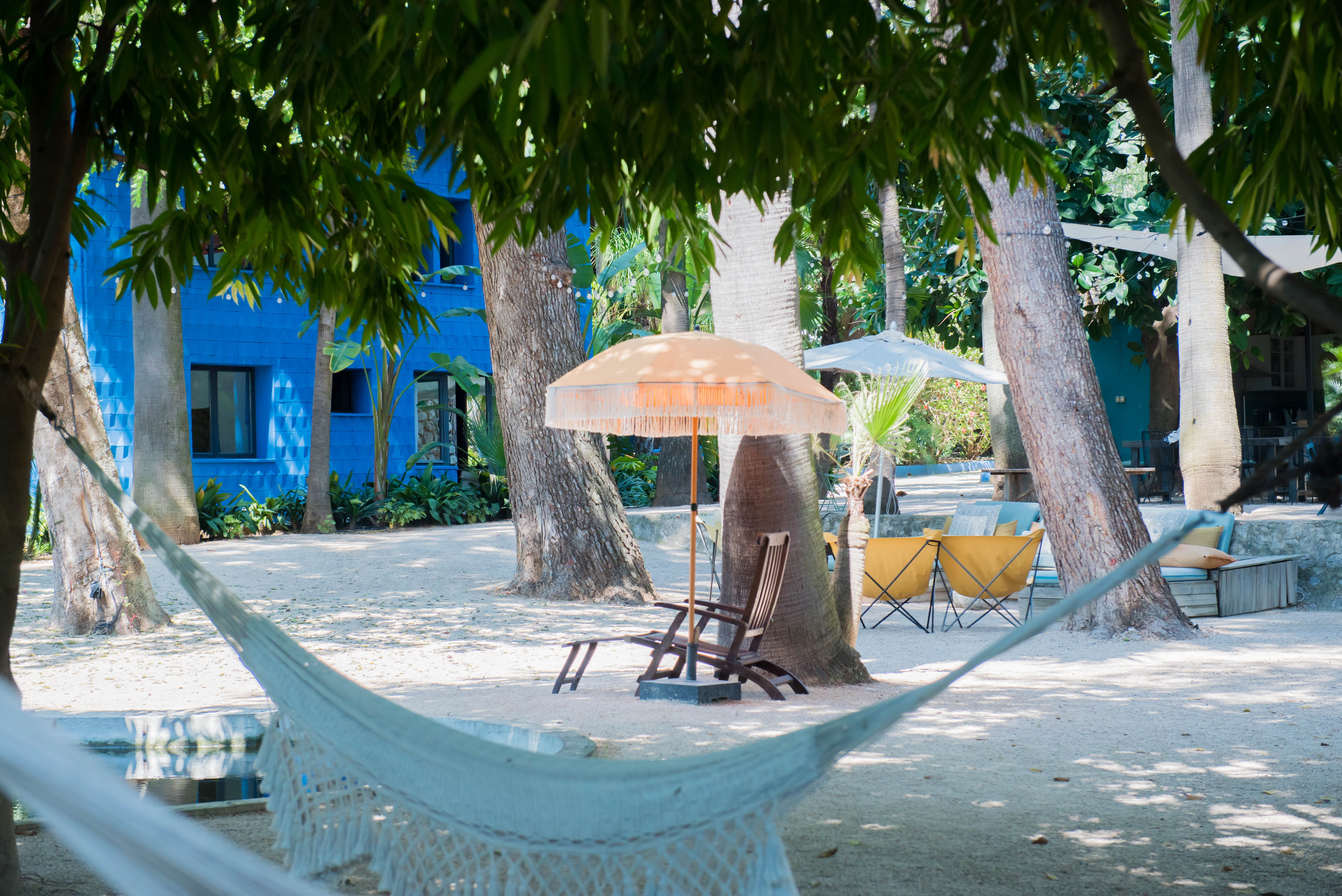 Espace de détente extérieur serein avec un hamac blanc tressé, ombragé par des arbres luxuriants. Un bâtiment bleu texturé et des sièges sous des parasols frangés sont visibles à l'arrière-plan.