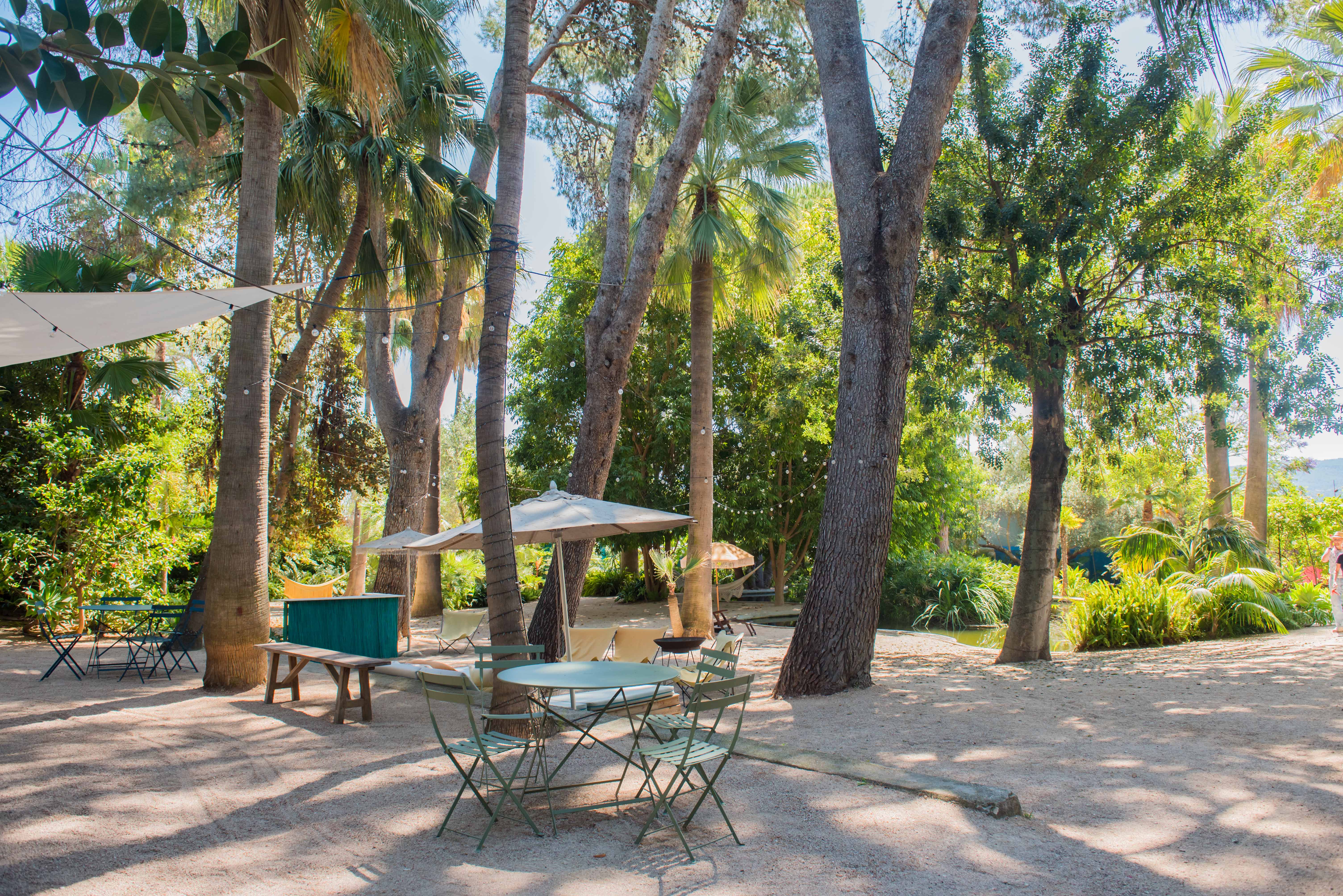 Espace extérieur aménagé avec tables et chaises en métal vert sur un sol de gravier. De grands palmiers et arbres matures procurent de l'ombre, agrémentés de parasols et guirlandes lumineuses dans un cadre tropical.