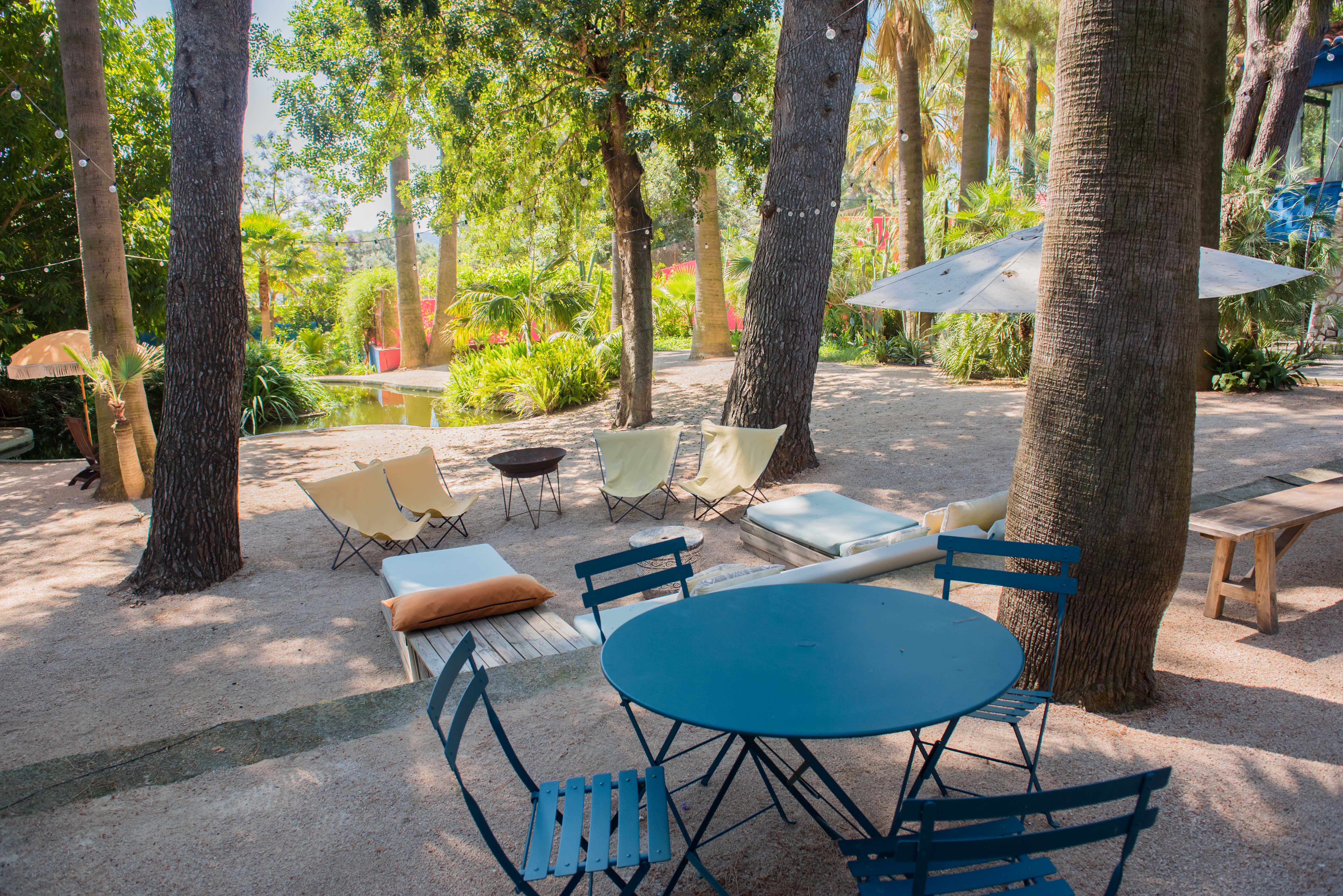Espace lounge extérieur avec ensemble bistro bleu et chaises en tissu sur gravier, ombragé par des arbres luxuriants avec guirlandes lumineuses, près d'un étang.
