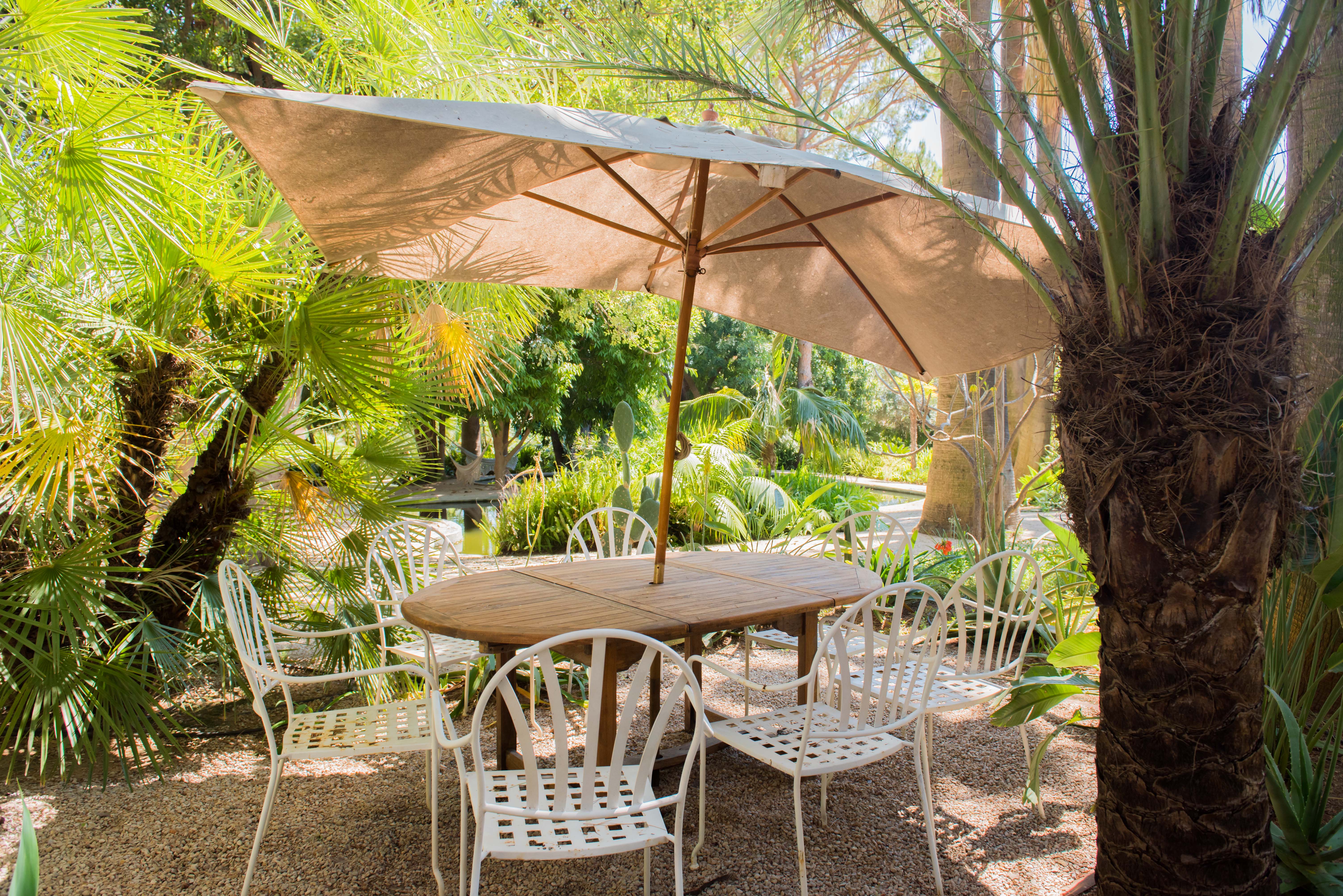 Espace repas extérieur avec table ronde en bois et six chaises en métal blanc sous un grand parasol beige. Entouré de palmiers tropicaux luxuriants et gravier, avec un étang visible en arrière-plan.