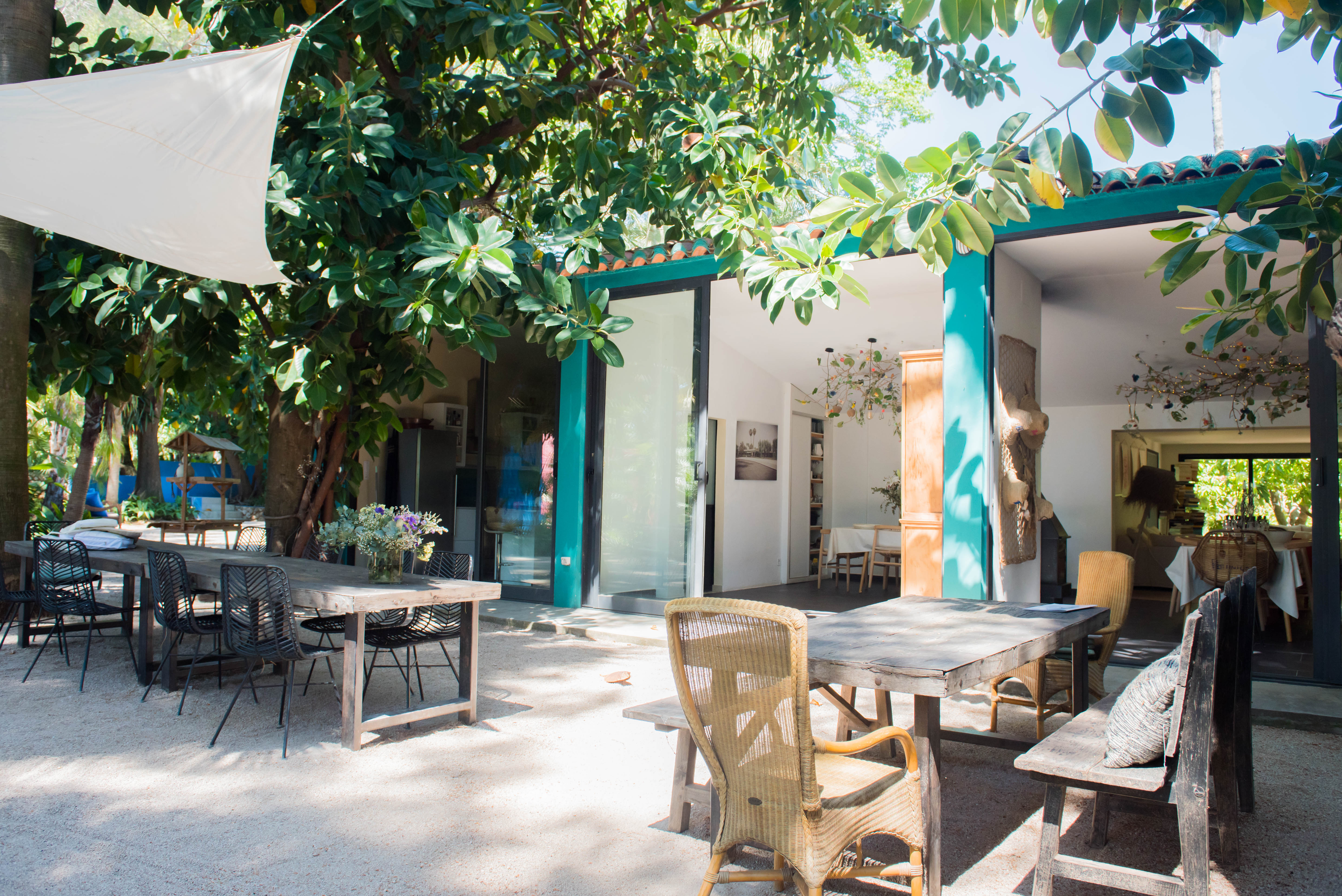 Espace repas extérieur avec tables en bois et sièges variés sous une voile d'ombrage blanche, encadré par une végétation tropicale luxuriante et relié à l'intérieur par des portes vitrées.