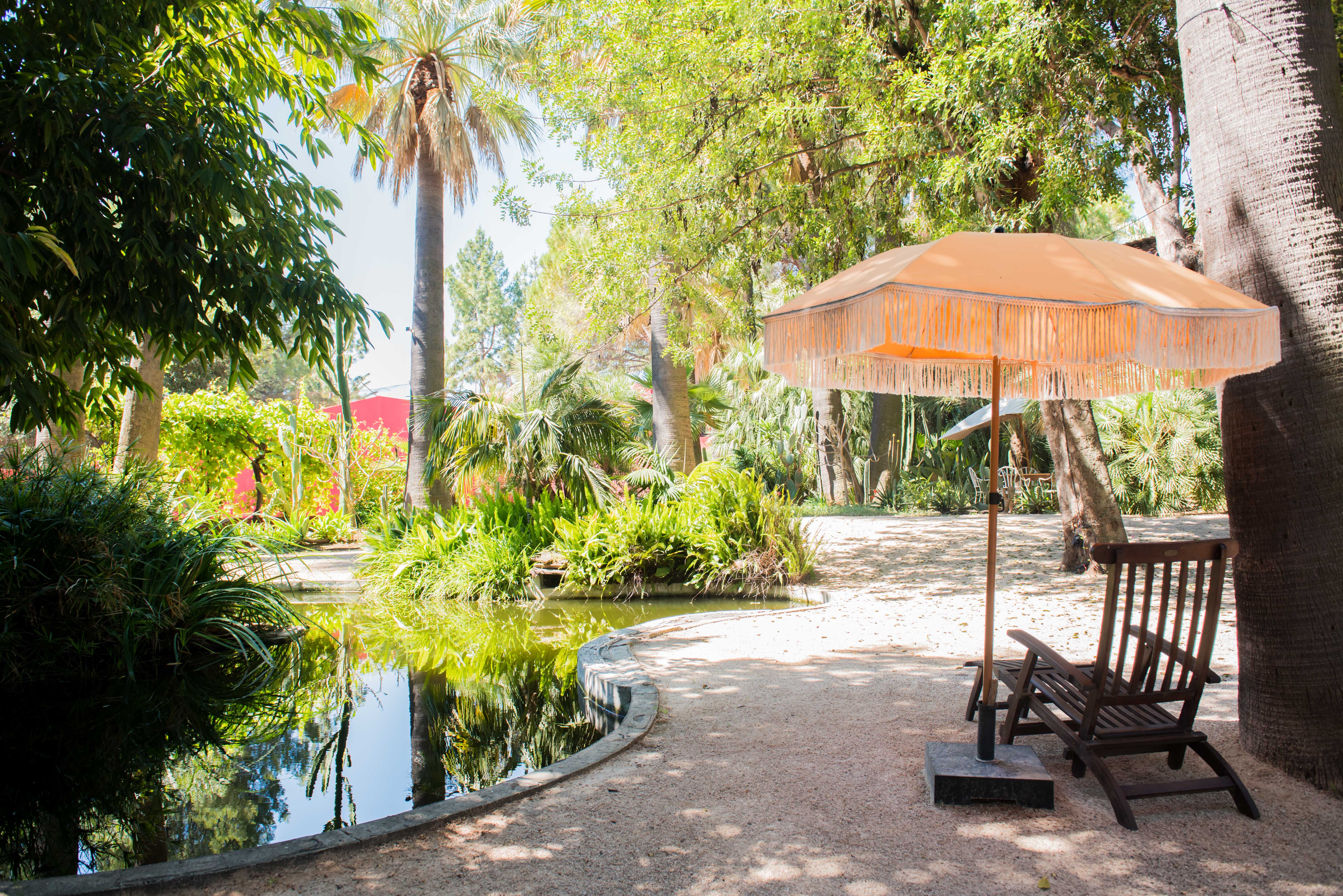 Étang paisible bordé d'une végétation tropicale luxuriante, avec un parasol orange à franges et une chaise longue en bois sur une allée de gravier.