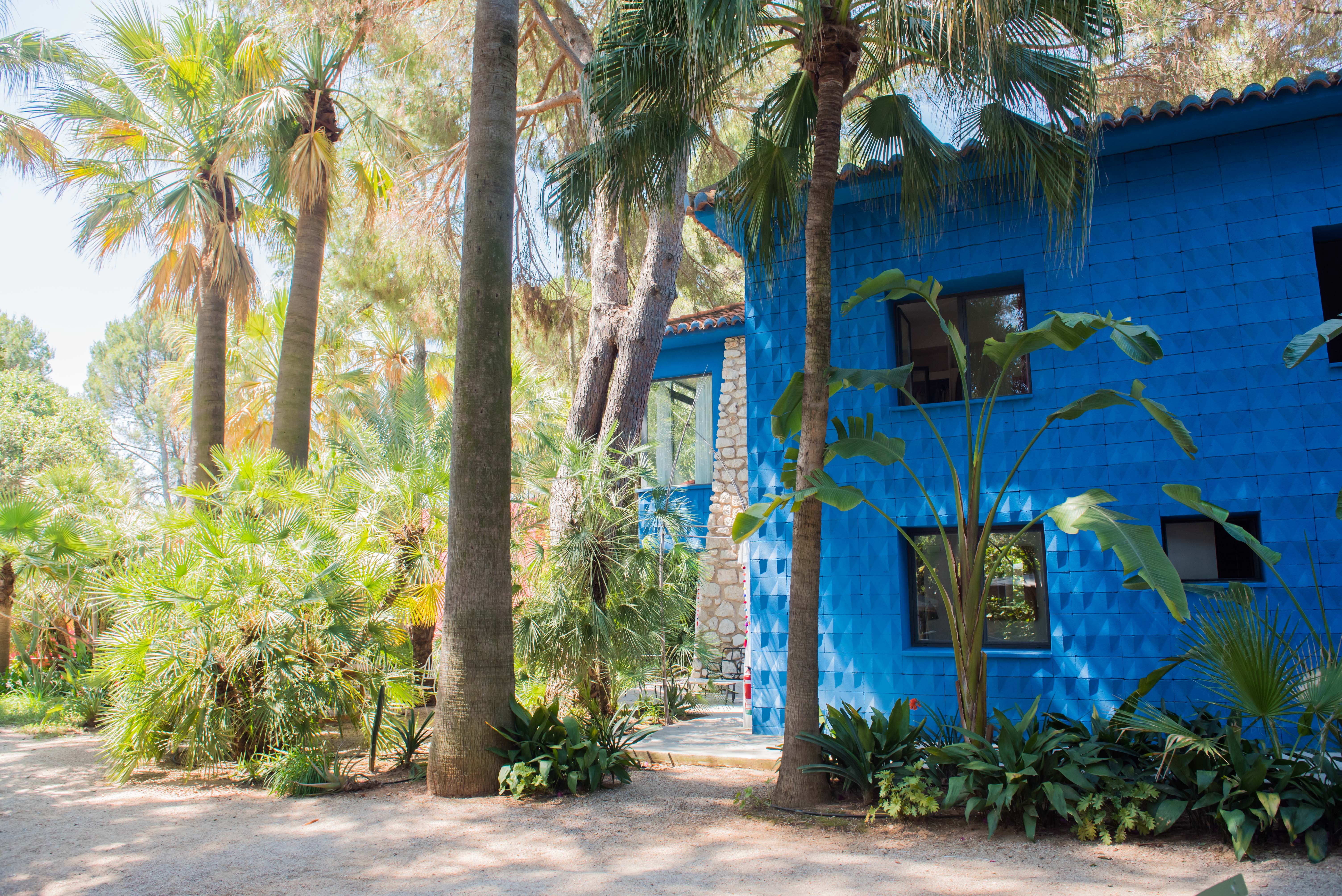 Façade bleue vibrante et texturée d'un bâtiment, entourée d'une végétation tropicale luxuriante. Fenêtres sombres et un toit de tuiles en terre cuite, avec une section en pierre apparente.
